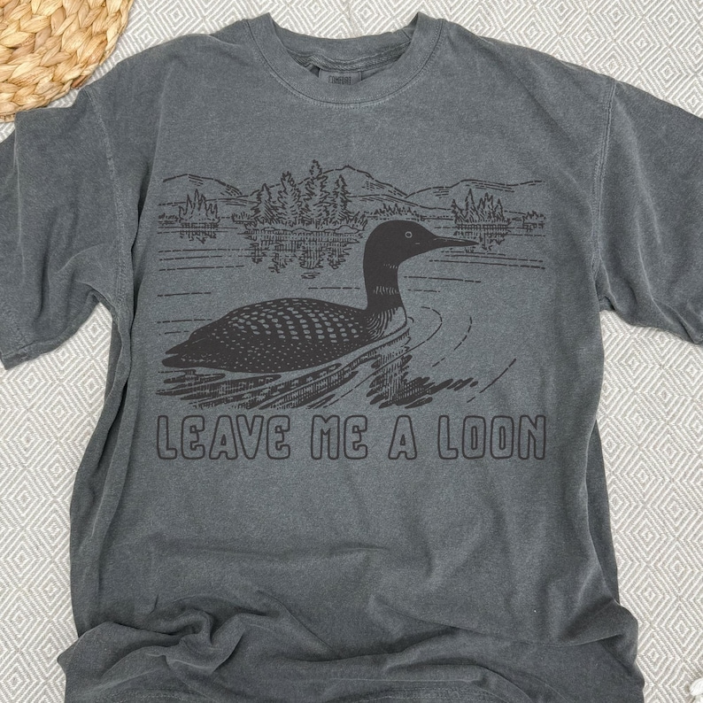 Puede incluir: Camiseta gris oscuro con un gr&aacute;fico negro de un somorgujo nadando en un lago, con monta&ntilde;as y &aacute;rboles al fondo. El texto "LEAVE ME A LOON" est&aacute; impreso debajo. La camiseta tiene mangas cortas.