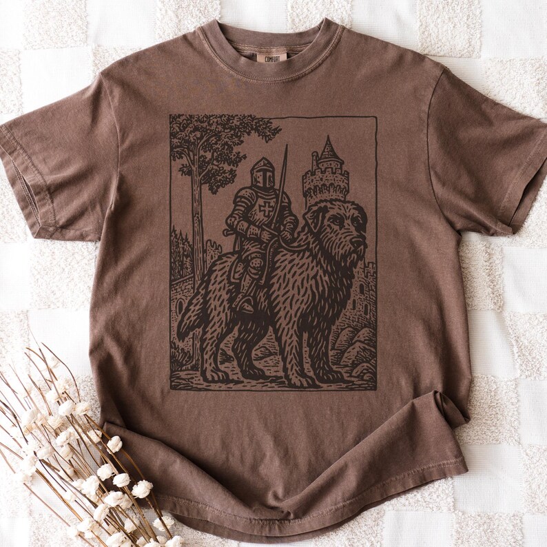 May include: Brown t-shirt featuring a black and white illustration of a knight riding a large dog, with a castle and tree in the background. The design has a vintage, woodcut style.