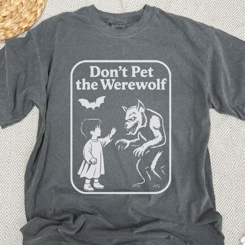 May include: Dark gray t-shirt with a white graphic that reads "Don't Pet the Werewolf." The graphic depicts a child reaching toward a werewolf, with a bat above them. The shirt is made of a soft material.