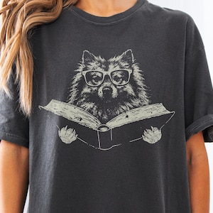 Może przedstawiać: Ciemnoszary t-shirt z krótkim rękawem z nadrukiem graficznym przedstawiającym puszystego psa w okularach z grubymi oprawkami, trzymającego otwartą książkę. Ilustracja wykonana jest w jasnym, złamanej bieli kolorze na ciemnej tkaninie.