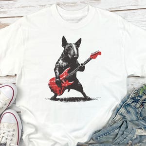 K&ouml;nnte beinhalten: Wei&szlig;es T-Shirt mit einer schwarzen Grafik eines Bullterriers, der eine rote E-Gitarre spielt. Der Hund ist in einer dynamischen Pose dargestellt und h&auml;lt die Gitarre. Das T-Shirt wird mit Jeans und wei&szlig;en Turnschuhen kombiniert.