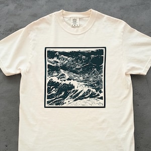 May include: Cream-colored t-shirt featuring a dark teal square graphic of ocean waves. The waves are depicted in a detailed, artistic style, creating a striking contrast against the shirt's neutral background. The shirt has short sleeves and a crew neck.