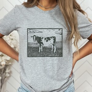 May include: A gray t-shirt featuring a black and white cow illustration within a rectangular frame. The cow stands in a field under a cloudy sky. The shirt is made of a soft material.