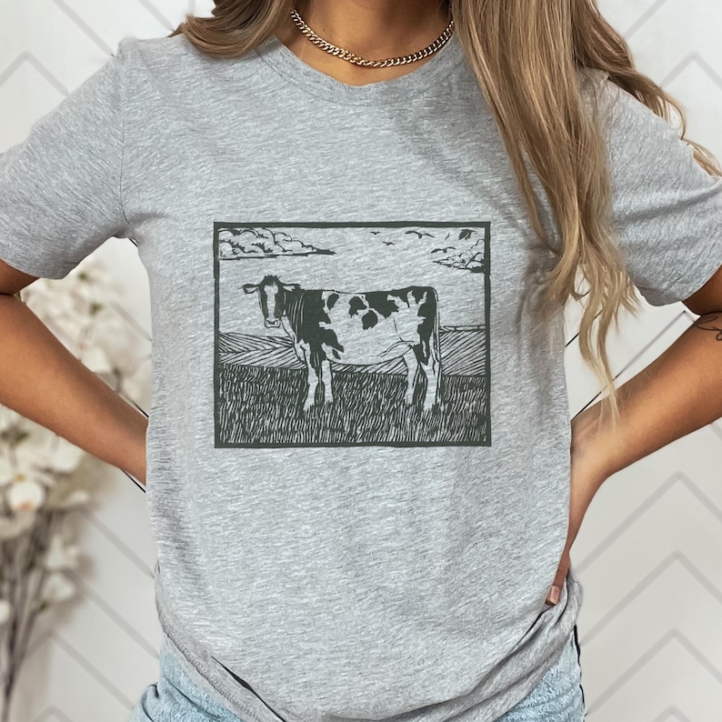 May include: A gray t-shirt featuring a black and white cow illustration within a rectangular frame. The cow stands in a field under a cloudy sky. The shirt is made of a soft material.