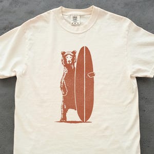 Puede incluir: Camiseta color crema con un oso pardo sosteniendo una tabla de surf. El oso está de pie, y la tabla está colocada verticalmente. El diseño es sencillo, perfecto para un estilo casual.