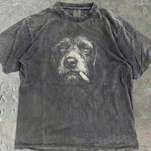 Könnte beinhalten: Dunkelgraues T-Shirt mit einem Grafikdruck eines Hundes mit Zigarette im Maul. Das Gesicht des Hundes ist weiß auf dem dunkelgrauen Stoff. Das T-Shirt hat einen Rundhalsausschnitt und kurze Ärmel. Der Stoff hat einen Wascheffekt.