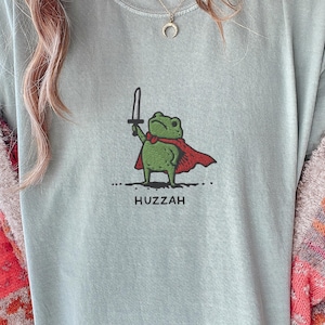 May include: A light blue t-shirt featuring a cartoon frog with a sword and a red cape. The frog is standing on a dotted line, with the word "Huzzah" printed below. The shirt is a casual style.