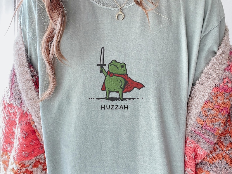 May include: A light blue t-shirt featuring a cartoon frog with a sword and a red cape. The frog is standing on a dotted line, with the word "Huzzah" printed below. The shirt is a casual style.