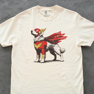 May include: Cream-colored t-shirt featuring a graphic of a dog dressed as a superhero. The dog wears a red mask, cape, and chest emblem with gold accents. The design has a vintage, distressed look.