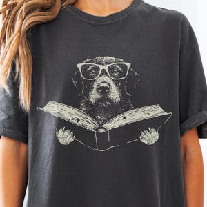 May include: A charcoal gray short-sleeve t-shirt featuring a graphic print of a black dog wearing white-rimmed glasses while holding an open book. The dog is centered on the chest, and the illustration is rendered in a light, off-white ink color.