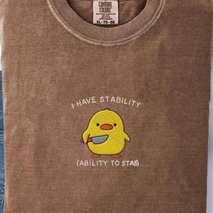 May include: Brown Comfort Colors t-shirt with the text "I HAVE STABILITY" and "(ABILITY TO STAB)". The shirt features an embroidered yellow chick holding a knife. The shirt is size XL-TG-EG.