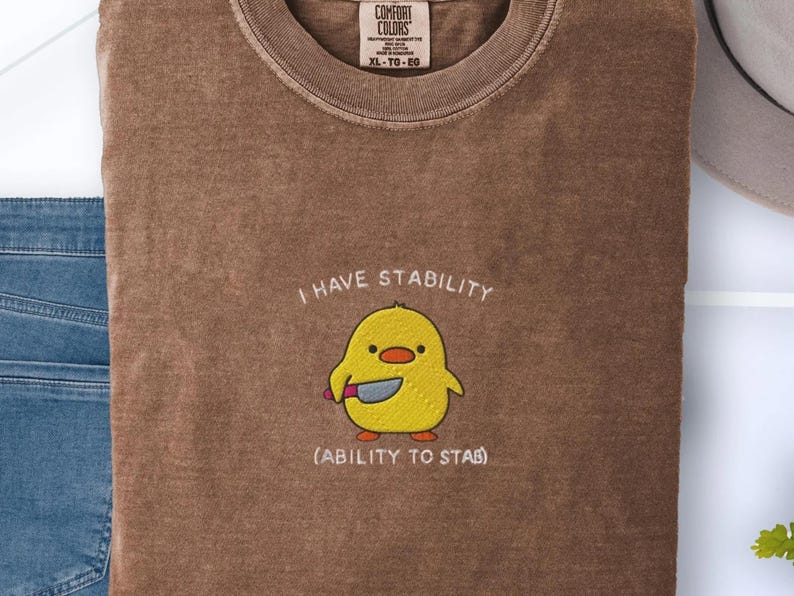 May include: Brown Comfort Colors t-shirt with the text "I HAVE STABILITY" and "(ABILITY TO STAB)". The shirt features an embroidered yellow chick holding a knife. The shirt is size XL-TG-EG.
