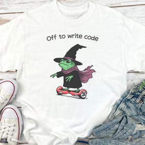 以下が含まれることがあります： 「Off to write code」の文字が入った白いTシャツ。緑色のカエルが魔女の帽子、スカーフ、マントを身につけ、赤いホバーボードに乗っているグラフィック。カエルは左を向いている。