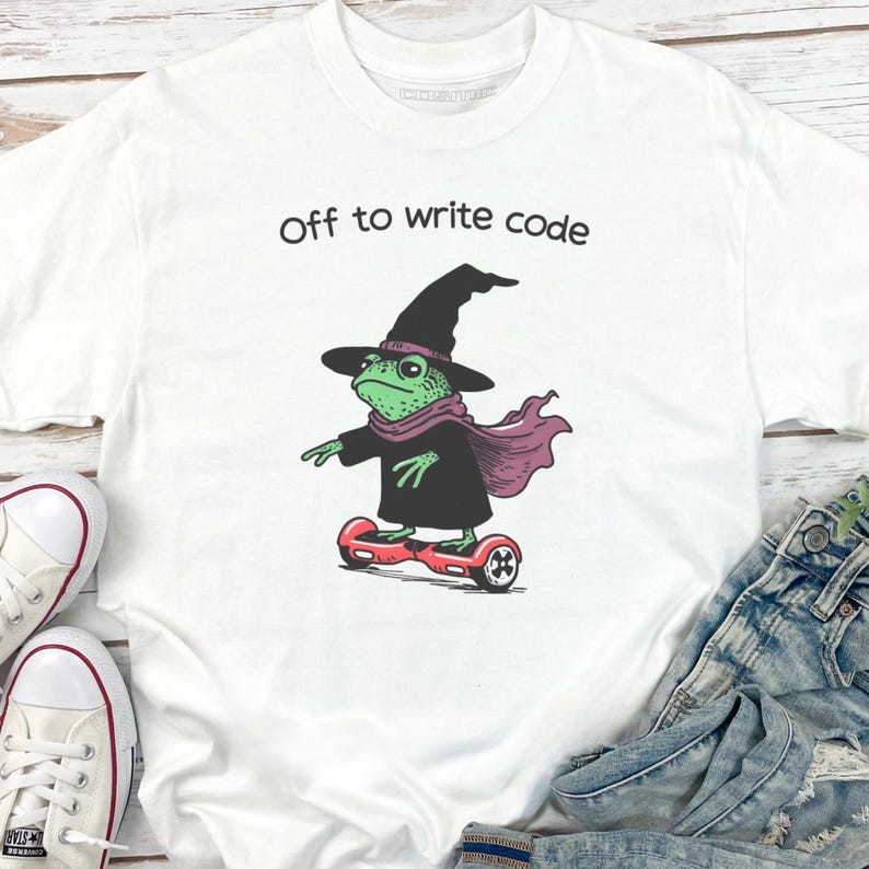 以下が含まれることがあります： 「Off to write code」の文字が入った白いTシャツ。緑色のカエルが魔女の帽子、スカーフ、マントを身につけ、赤いホバーボードに乗っているグラフィック。カエルは左を向いている。