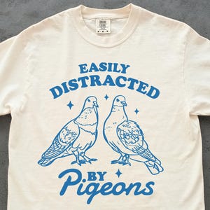 May include: Off-white t-shirt with the phrase "EASILY DISTRACTED BY PIGEONS" in blue. The design includes two blue pigeon illustrations and four blue star shapes.