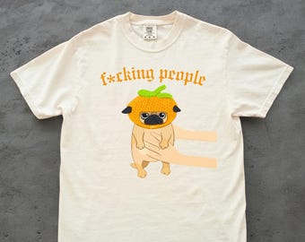 Könnte beinhalten: Cremefarbenes T-Shirt mit einem Cartoon-Mops, der einen orangefarbenen Kürbishut trägt. Der Text "f*cking people" ist in orangefarbenen gotischen Buchstaben über dem Mops aufgedruckt. Der Mops wird von zwei Cartoon-Händen gehalten.