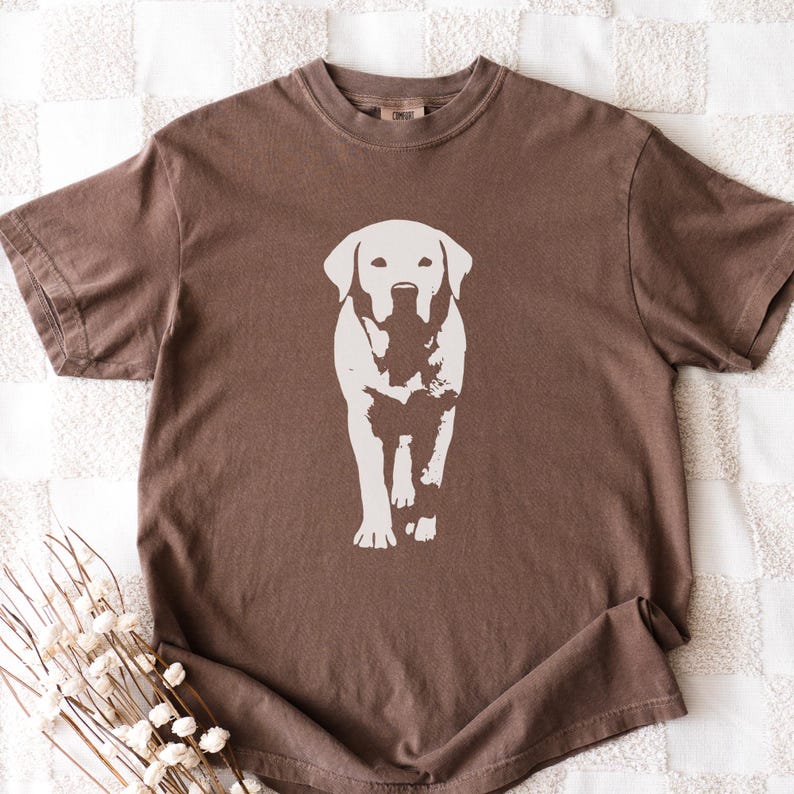 May include: A brown t-shirt featuring a white graphic of a Labrador Retriever dog. The dog is centered on the shirt, facing forward. The shirt has short sleeves and a crew neck.