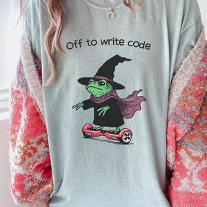 以下が含まれることがあります： 「Off to write code」の文字が入った水色のTシャツ。魔女の帽子、スカーフ、マントを着た緑色のカエルが赤いホバーボードに乗っているデザイン。カエルがメインのグラフィックです。