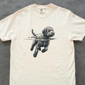 Könnte beinhalten: Cremefarbenes T-Shirt mit einer Schwarz-Weiß-Illustration eines Hundes, der im Wasser schwimmt. Der Hund mit lockigem Fell ist mittig auf dem Shirt abgebildet.