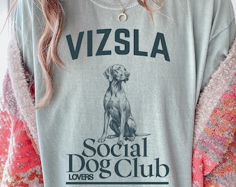 May include: A pale blue t-shirt featuring the word "VIZSLA" above a detailed illustration of a Vizsla dog. Below the dog is the text "Social Dog Club Lovers". The shirt has a relaxed fit.