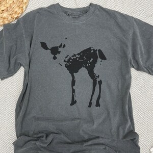 May include: Dark gray t-shirt featuring a black silhouette of a deer. The deer has a spotted pattern and is standing. The shirt is made of a soft material and has a crew neck.