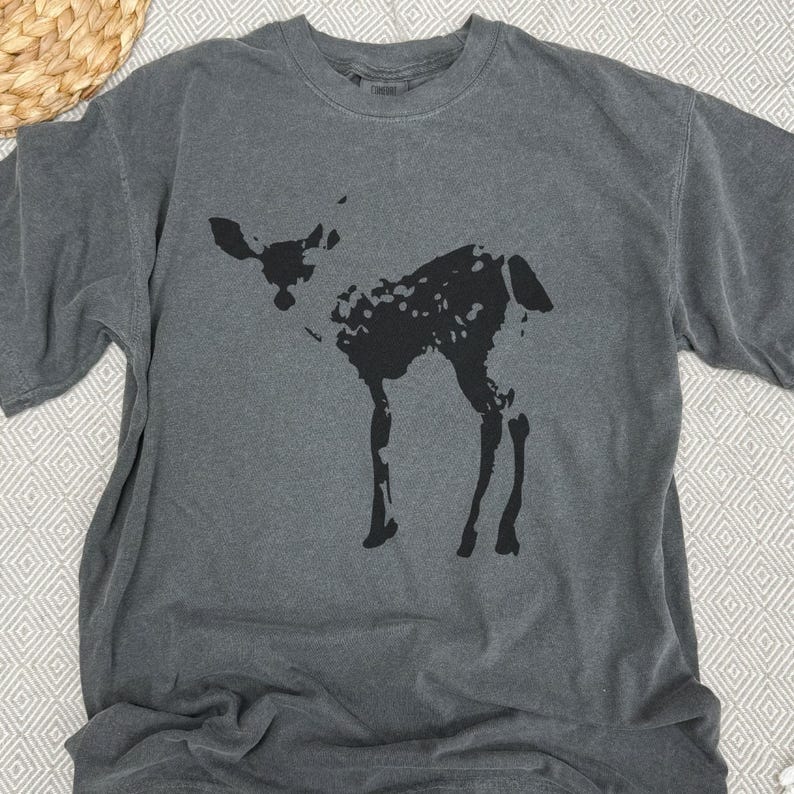 May include: Dark gray t-shirt featuring a black silhouette of a deer. The deer has a spotted pattern and is standing. The shirt is made of a soft material and has a crew neck.