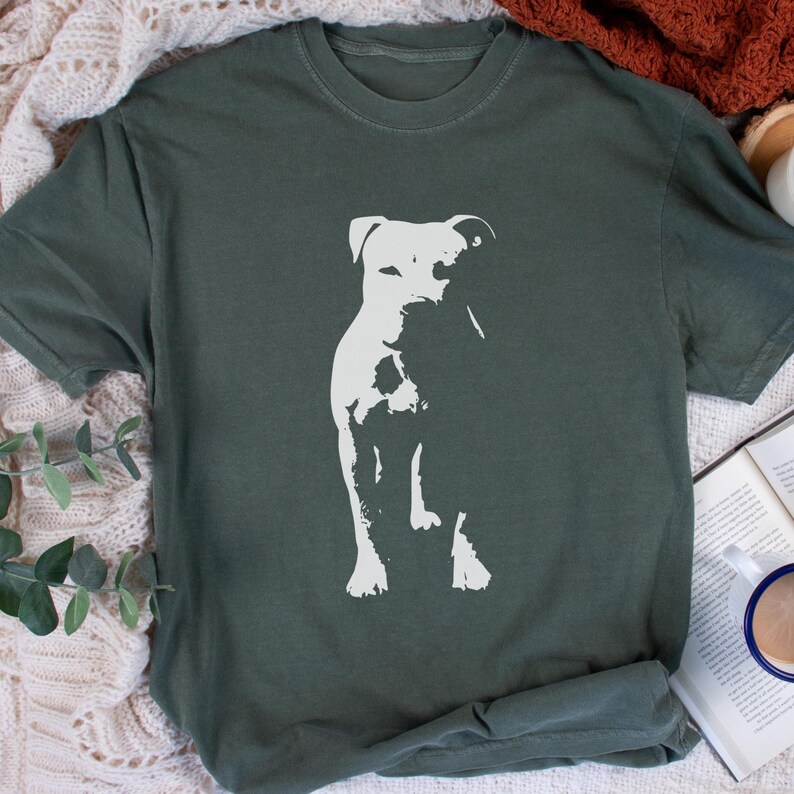 Pode incluir: T-shirt verde escuro com uma silhueta branca de um c&atilde;o. O c&atilde;o &eacute; mostrado em p&eacute;, com seus tra&ccedil;os sutilmente delineados. A camisa &eacute; feita de um material macio e tem um corte folgado.
