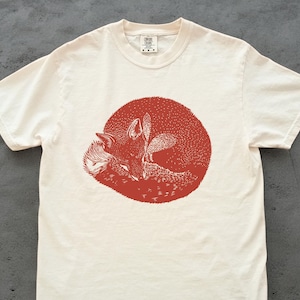 Peut inclure: T-shirt blanc cassé avec une illustration rouge d'un renard enroulé dans un cercle. Le motif du renard est détaillé avec des points, créant un aspect texturé. Le t-shirt a des manches courtes et un col rond.