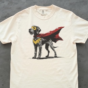 Puede incluir: Camiseta color crema con una ilustración de un perro superhéroe. El perro lleva una capa roja, una máscara amarilla y un emblema dorado en el pecho. El diseño está centrado en la camiseta, creando un diseño divertido y atractivo.