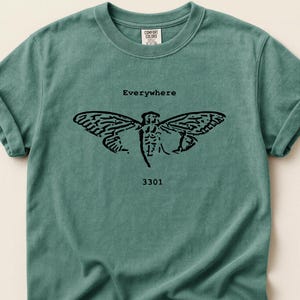 May include: A teal Comfort Colors t-shirt featuring a black cicada graphic with the word "Everywhere" above it and the number "3301" below. The shirt has rolled-up sleeves and a crew neck.