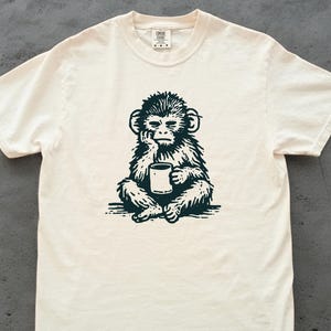 May include: A cream-colored short-sleeve t-shirt featuring a dark green graphic print of a monkey sitting and holding a mug. The monkey has its hand resting on its chin in a contemplative pose. The shirt has a crew neckline and a brand label at the neck.