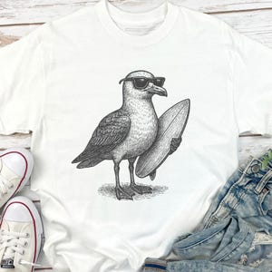 K&ouml;nnte beinhalten: Ein wei&szlig;es T-Shirt mit einer schwarz-wei&szlig;en Strichzeichnung einer M&ouml;we, die eine Sonnenbrille tr&auml;gt und ein Surfbrett unter dem Fl&uuml;gel h&auml;lt. Die Grafik ist mittig auf der Brust platziert und zeigt detaillierte Federstrukturen und Schattierungen.