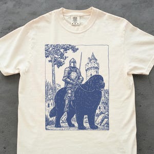 May include: Cream-colored t-shirt featuring a blue illustration of a knight in armor riding a large dog, with a castle in the background. The design is detailed and reminiscent of a medieval theme.