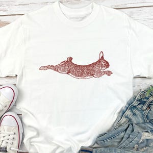 Puede incluir: Camiseta blanca con una ilustraci&oacute;n marr&oacute;n rojiza de un conejo corriendo. El conejo est&aacute; representado de forma detallada y estilizada. La camiseta se combina con vaqueros azules y zapatillas blancas con detalles rojos.