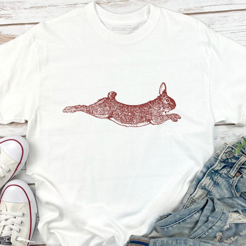 Puede incluir: Camiseta blanca con una ilustraci&oacute;n marr&oacute;n rojiza de un conejo corriendo. El conejo est&aacute; representado de forma detallada y estilizada. La camiseta se combina con vaqueros azules y zapatillas blancas con detalles rojos.