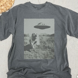 May include: A dark gray t-shirt with a black and white graphic print of a dog looking up at a flying saucer. The graphic is centered on the shirt.
