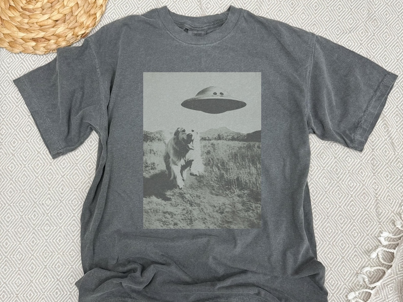 May include: A dark gray t-shirt with a black and white graphic print of a dog looking up at a flying saucer. The graphic is centered on the shirt.