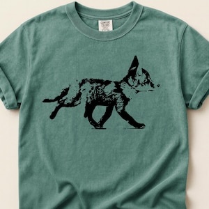 Puede incluir: Una camiseta de color verde azulado con una silueta negra de un zorro corriendo. El gráfico del zorro es detallado, con textura de pelaje visible. La camiseta tiene mangas enrolladas y cuello redondo, hecha de un material suave.