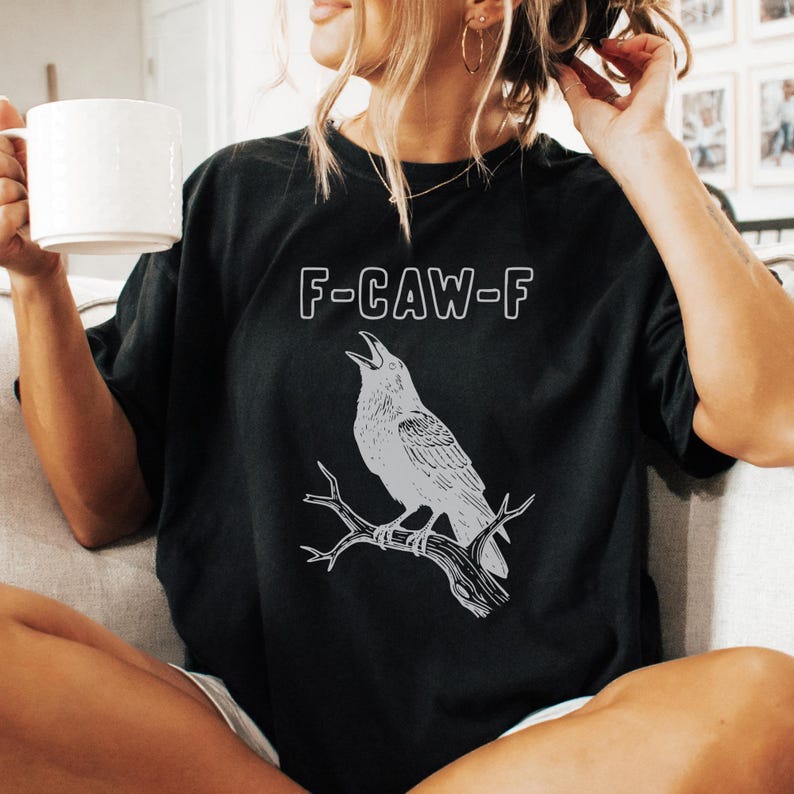 May include: Black t-shirt with a gray illustration of a crow perched on a branch, with the text "F-CAW-F" above the bird. The design is simple and graphic, suitable for casual wear.