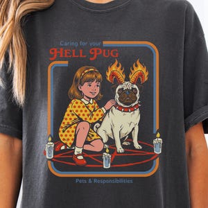 May include: Dark gray t-shirt featuring a graphic of a girl and a pug with flames on its head, surrounded by candles and a pentagram. The text reads "Caring for your Hell Pug" and "Pets & Responsibilities."
