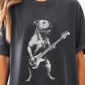 Pode incluir: Camiseta cinza escuro com uma ilustração em preto e branco de um cão tocando baixo. O cão está em pé e parece estar cantando. O design tem um visual vintage.