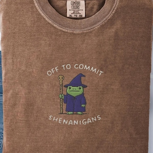 Może przedstawiać: Brązowa koszulka z białym napisem "OFF TO COMMIT SHENANIGANS". Koszulka przedstawia haftowaną kreskówkową żabę w czapce i szacie czarodzieja, trzymającą laskę. Koszulka to Comfort Colors XL-TG-EG.