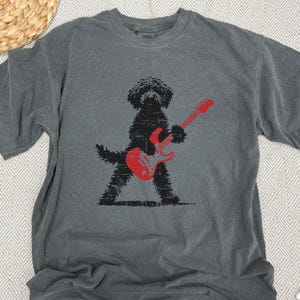 May include: Gray t-shirt featuring a black dog playing a red electric guitar. The dog is silhouetted with a textured design. The shirt has a crew neck and short sleeves. The background is a neutral color.
