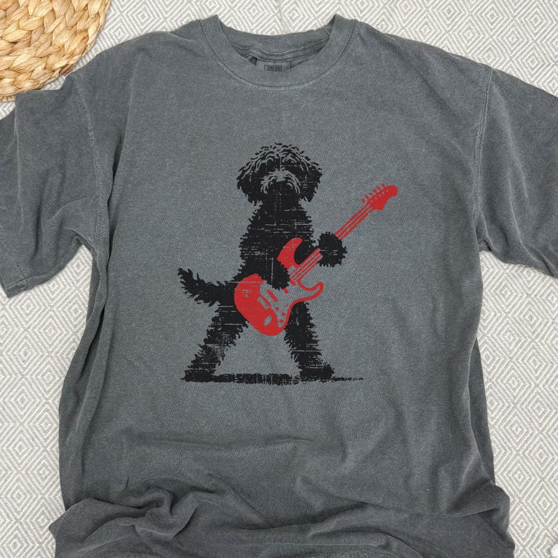 May include: Gray t-shirt featuring a black dog playing a red electric guitar. The dog is silhouetted with a textured design. The shirt has a crew neck and short sleeves. The background is a neutral color.