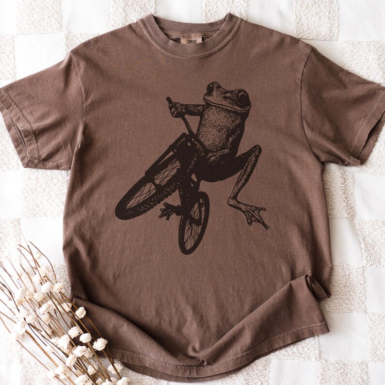 Pu&ograve; includere: T-shirt marrone con una grafica nera di una rana in bicicletta. La rana e la bicicletta sono dettagliate. La maglietta ha un girocollo e maniche corte. Il tessuto sembra morbido. Il design &egrave; giocoso e fantasioso.