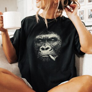 May include: Black t-shirt featuring a detailed, grayscale illustration of a gorilla smoking a cigarette. The gorilla's face is the focal point, with a serious expression. The shirt is a relaxed fit, suitable for casual wear.