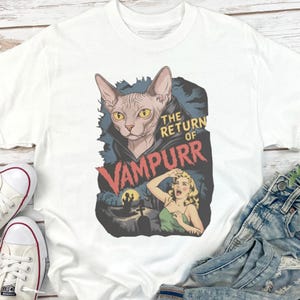 Peut inclure: T-shirt blanc avec un motif graphique. Le motif repr&eacute;sente un chat Sphynx portant une cape de vampire, le texte "The Return of Vampurr", une femme qui crie, un cimeti&egrave;re et une pleine lune.