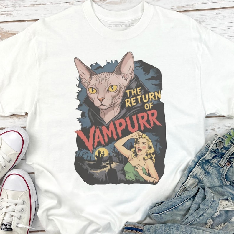 Peut inclure: T-shirt blanc avec un motif graphique. Le motif repr&eacute;sente un chat Sphynx portant une cape de vampire, le texte "The Return of Vampurr", une femme qui crie, un cimeti&egrave;re et une pleine lune.