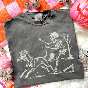May include: A charcoal gray crewneck sweatshirt featuring a graphic print of a skeleton reaching toward a dog that is holding a bone in its mouth. The design includes a tombstone, a bare tree, and a detached bone on the ground. The shirt has a Comfort Colors label.