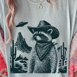 May include: A light blue t-shirt with a dark teal graphic of a raccoon in a cowboy hat and bandana, set in a desert scene with cacti and mountains. A UFO is also present. The design is detailed.
